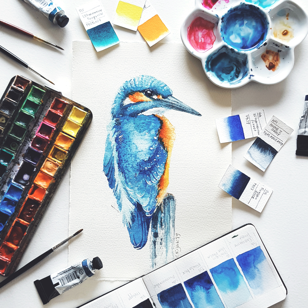 Akwarela zimorodka w otoczeniu akcesoriów malarskich. Farby akwarelowe i pędzelki.